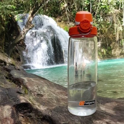 24oz OTF Bottle Clear with Orange Cap sitting on a rock in front of a waterfall