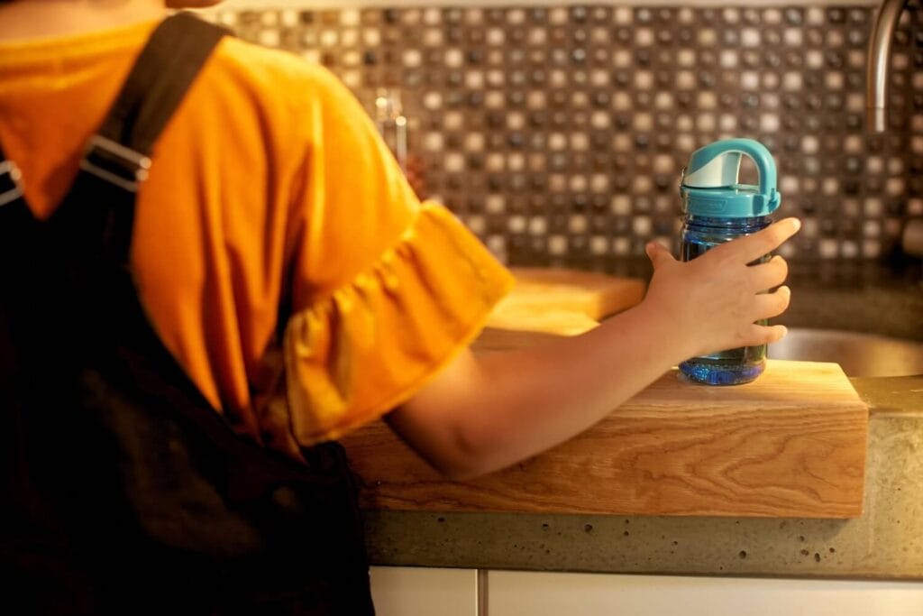 The image features a young child, visible from the back, holding a blue Nalgene water bottle. The child is dressed in a bright orange shirt and a black apron, standing at a kitchen counter. The background has a decorative tile wall, enhancing the cozy, domestic setting. The scene captures a moment of the child either preparing to fill up the bottle or having just filled it at the kitchen sink.