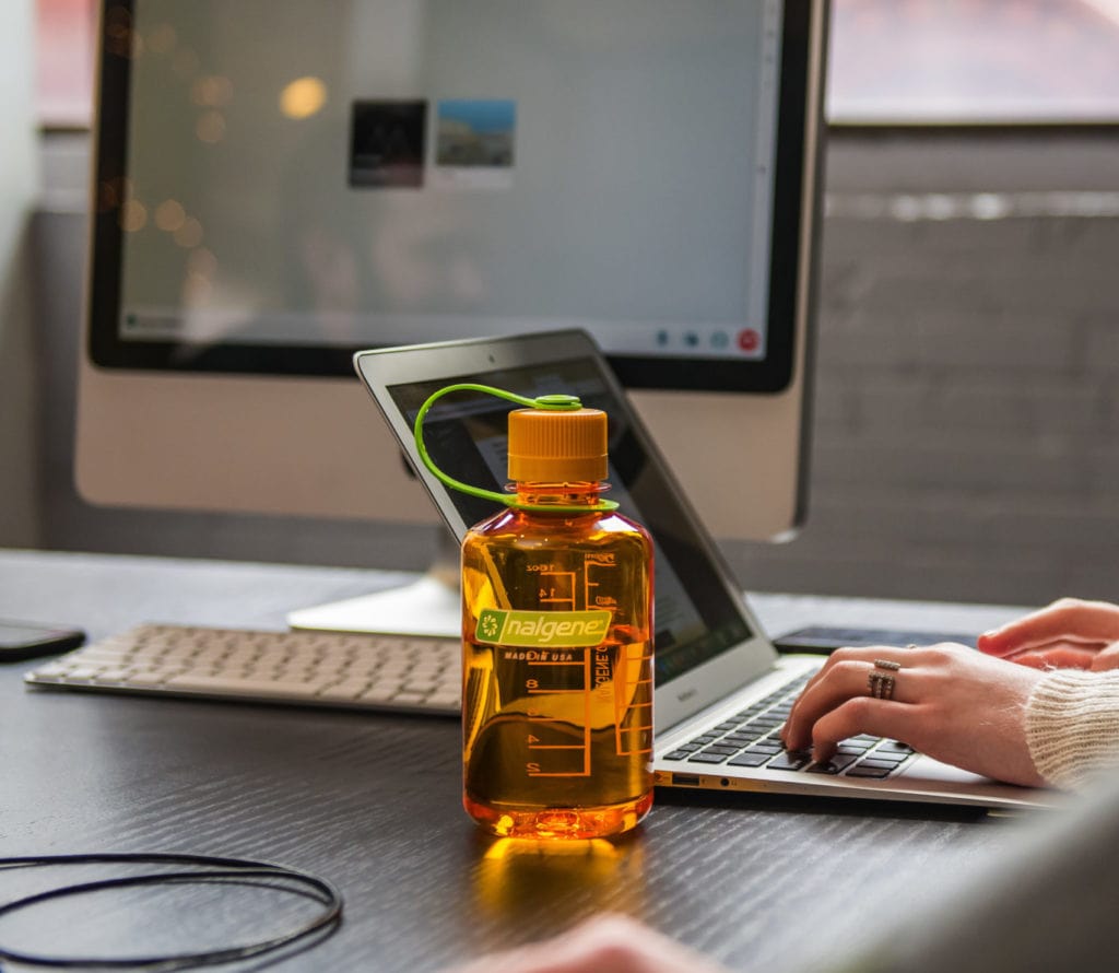 An orange Nalgene water bottle on a desk in front of a laptop and desktop computer.