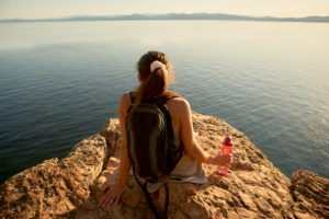 Woman with a backpack sits on a rocky cliff overlooking a calm lake, holding a water bottle.