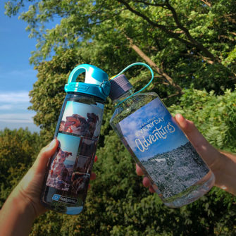 Hands holding up two customized Nalgene bottles. The bottle on the right is a 32oz Narrow Mouth Tritan Water Bottle with a landscape photograph, and the image on the left is a 24oz On-The-Fly Lock-Top Bottle with a collage of dog photos.