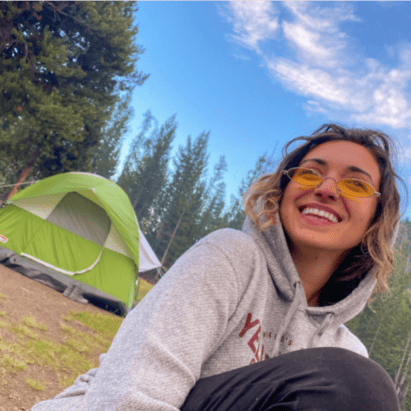 Michelle wearing yellow sunglases, sitting in front of a green tent set up in the forest