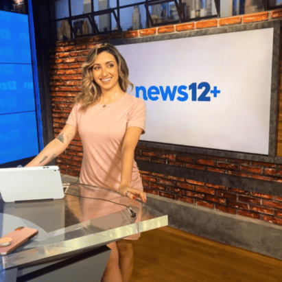 Michelle standing at a desk with the news12+ sign on a brick wall behind her