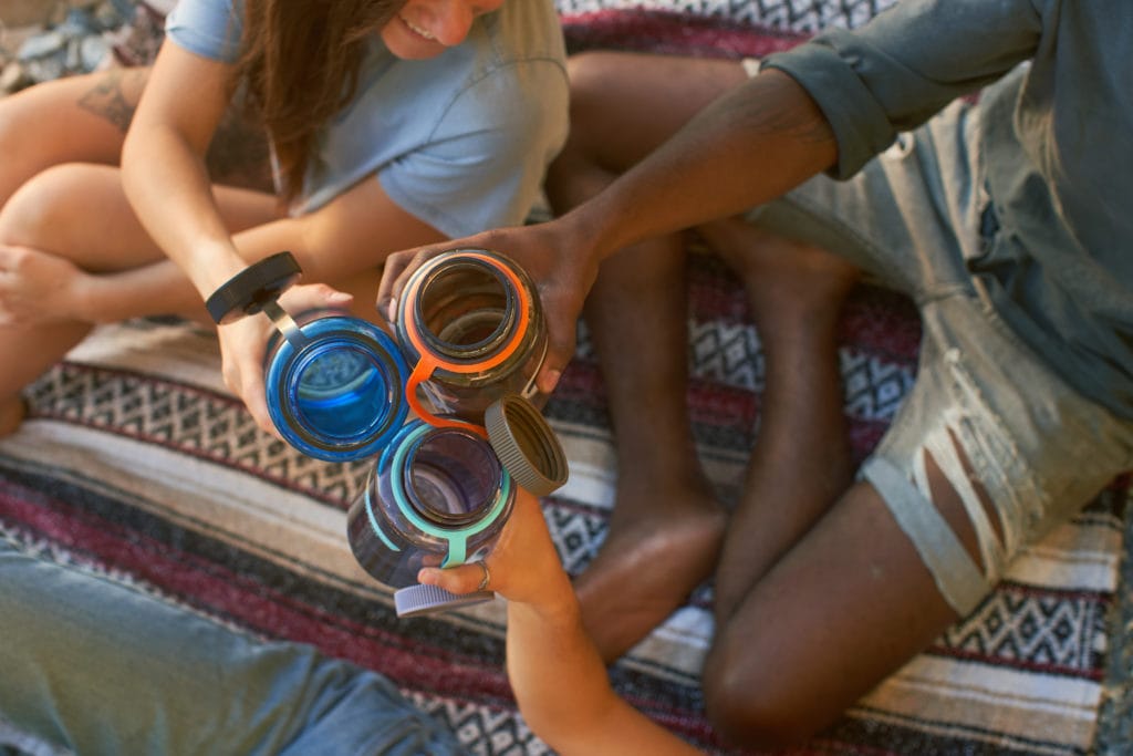 Three people sitting on a picnic blanket and holding up their Wide Mouth Nalgene bottles in a "cheers" motion