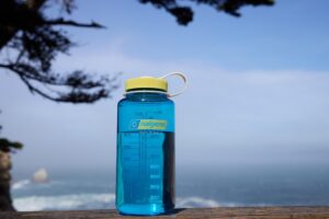 A blue Nalgene water bottle on a wooden surface with the ocean and trees in the background.