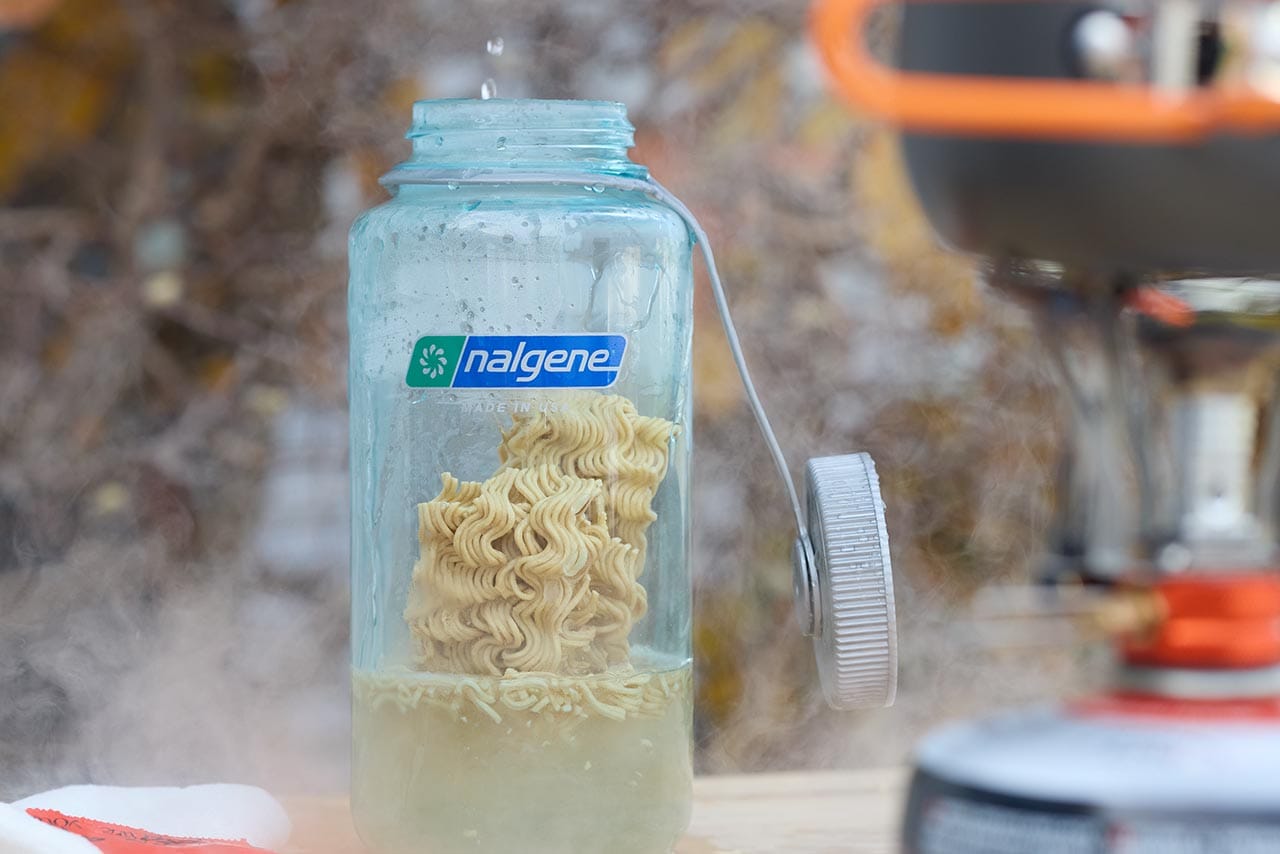 A Nalgene bottle filled with instant ramen and nalgene boiling water steams beside a portable stove, ready for a quick trail-side meal.