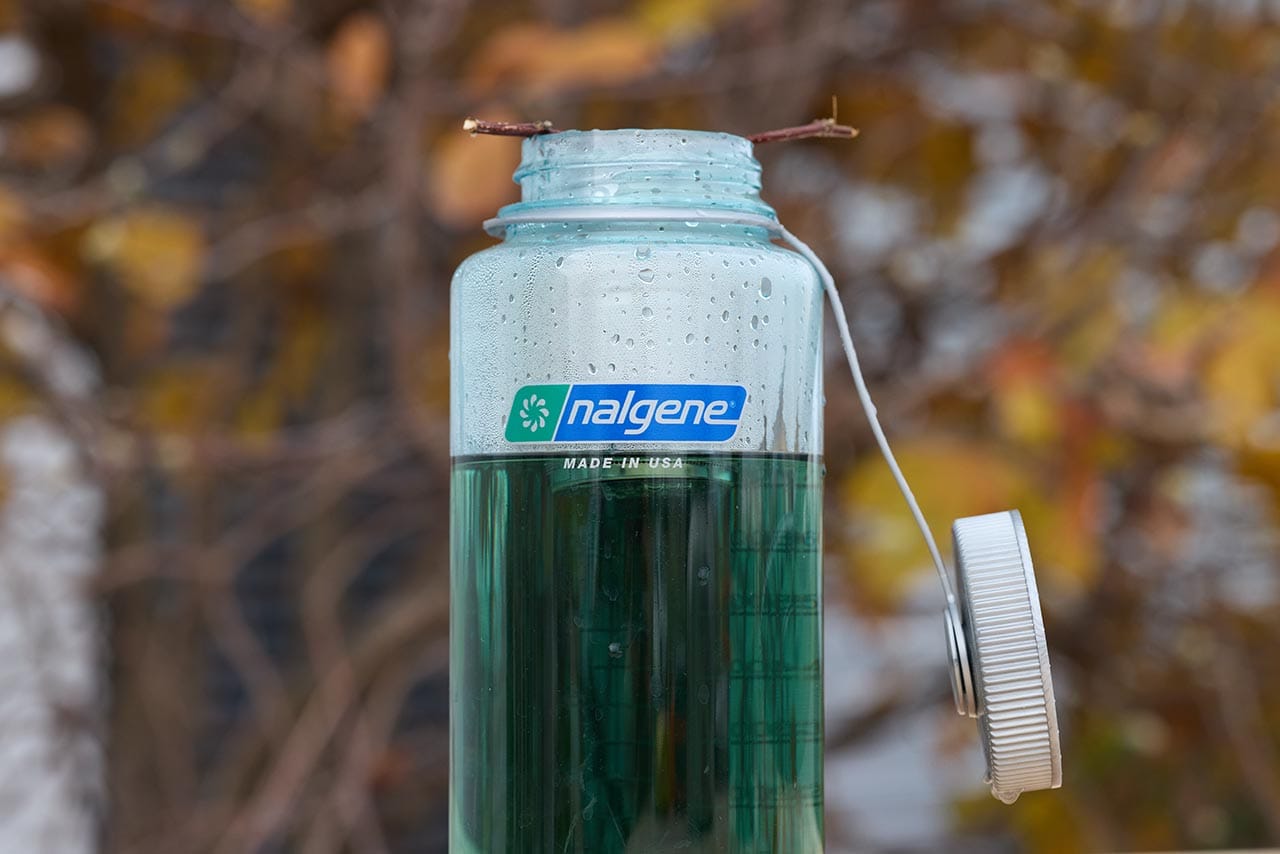 A Nalgene water bottle, known for handling boiling water, stands open outdoors filled with green liquid, autumn leaves in the background.