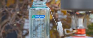 Clear Nalgene water bottle on a table with dry ramen noodles and a camping stove nearby, ready for boiling water outdoors.